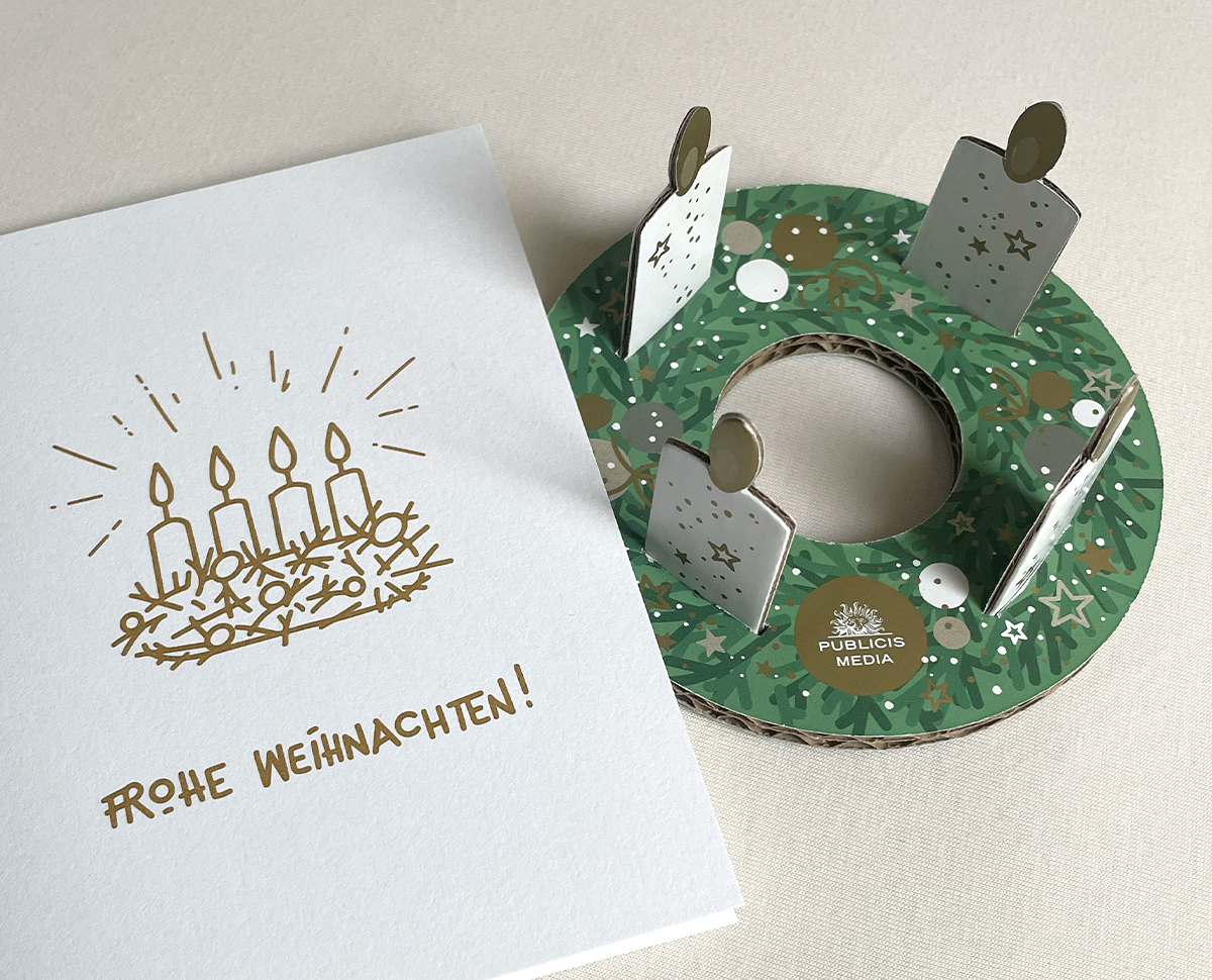 Foto von Weihnachtskarte (links) und Adventkranz aus Karton (rechts) für Publicis Media Austria. Cover der Weihnachtskarte (angeschnitten) mit Illustration (Strichgrafik, Outline in Gold) eines Adventkranzes. Darunter steht handschriftlich in Versalien "Frohe Weihnachten!" Adventkranz aus Karton mit Kerzen zum Einstecken. Copyright © 2023 Olivia Weise, Design & Illustration